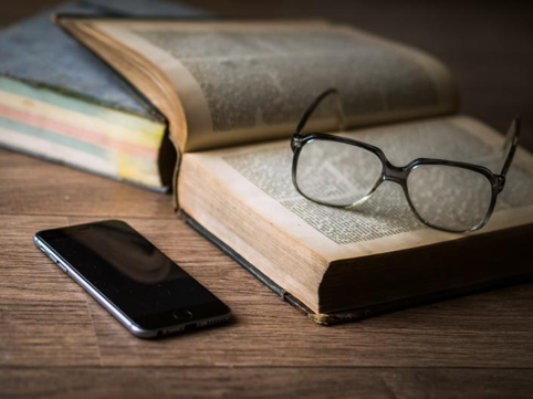 Image of a black pair of glasses resting on an opened book, which is resting on a wooden table with a mobile phone next to it.