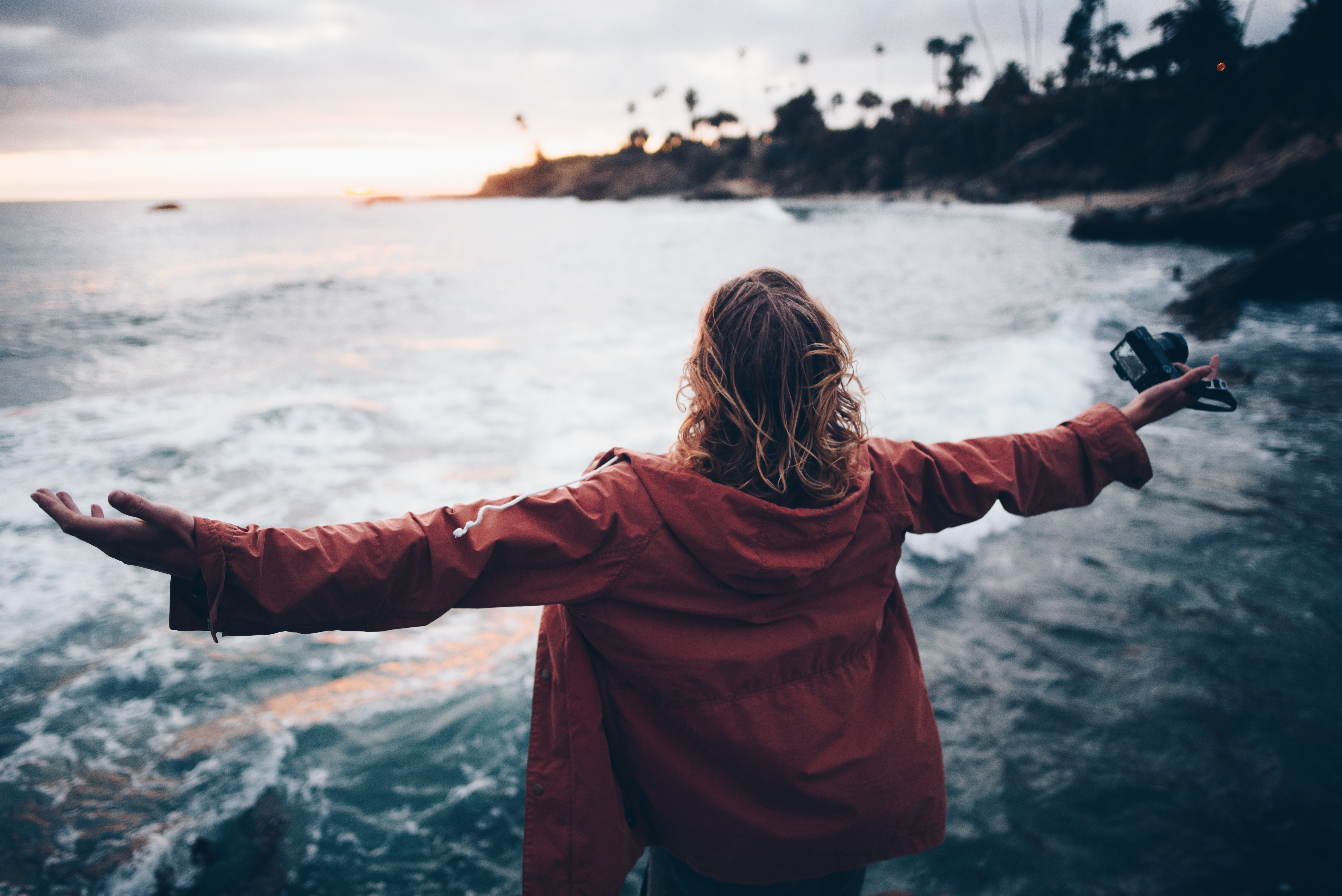 Image of a woman with blonde curly hair, wearing a red fleece, looking out over a long stretch of water, with her arms stretched out and only her back showing as she portrays a "freelance lifestyle."