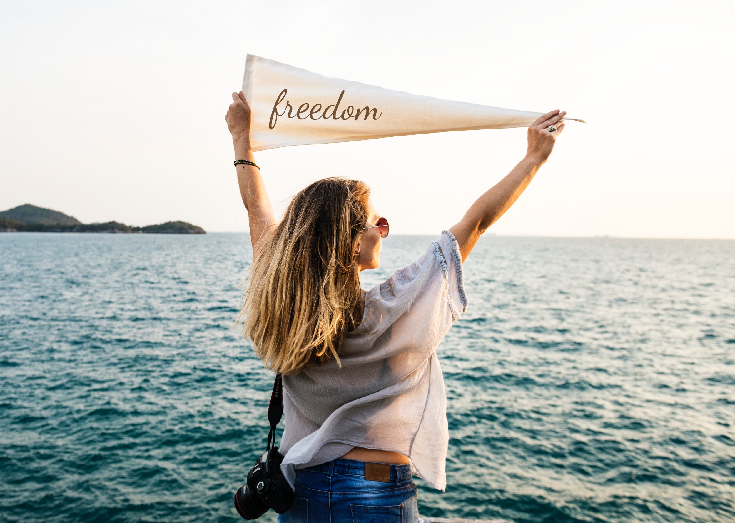 Image of a woman with blond hair, wearing sunglasses, blue jeans, and skin tight white top. She is standing in the ocean up to her waist, looking at the horizon with her back to us and holding a white banner with the word "freedom" written on it.