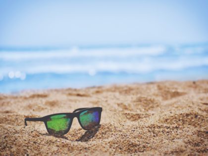 Image of a pair of sunglasses on a sandy beach with water as a background.
