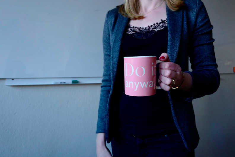 A woman wearing a dark outfit and standing in a light blue room; holding a large pink cup in her left hand.