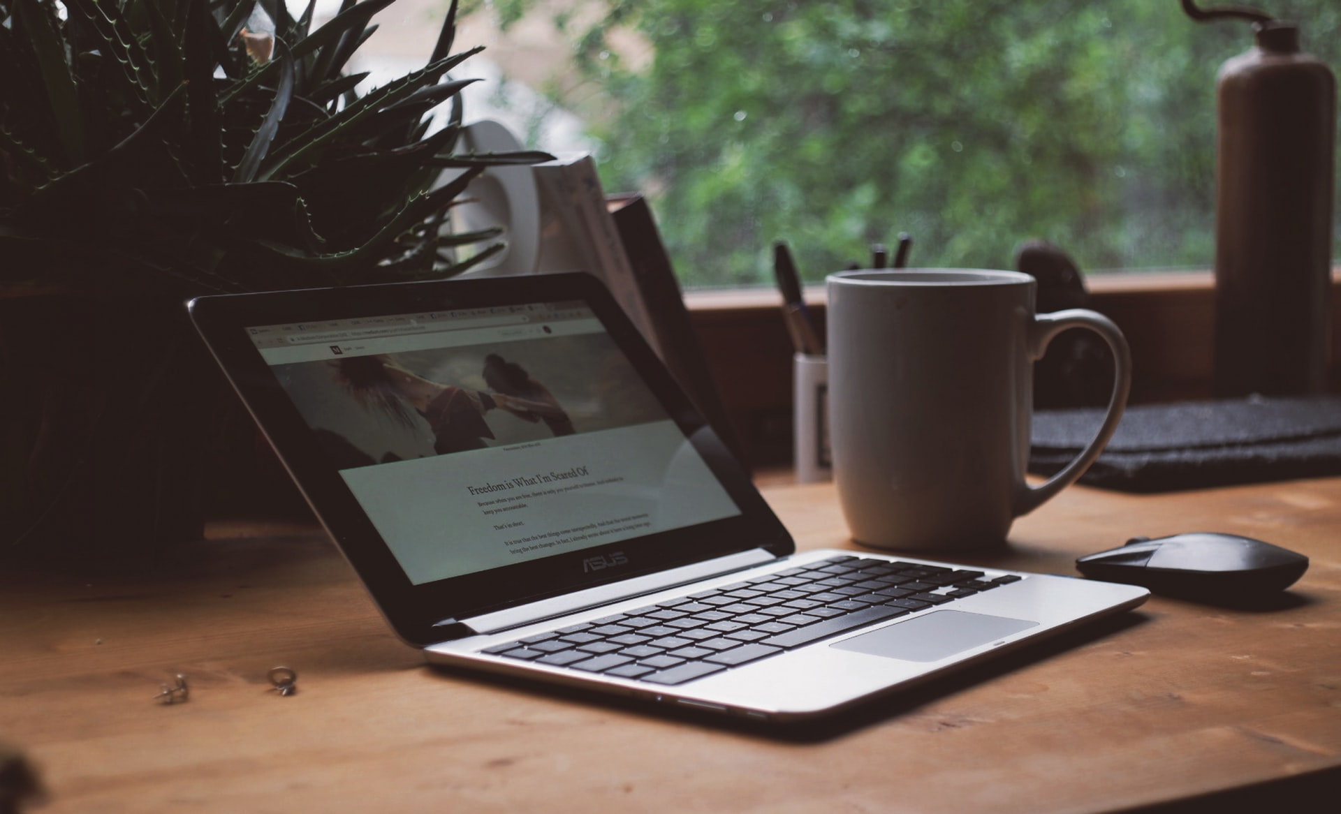 An open laptop on a desk with a coffee cup next to it and a window nearby.