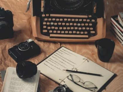 Image of an old typewriter, pen, open notebook with writing in it, glasses and a camera.
