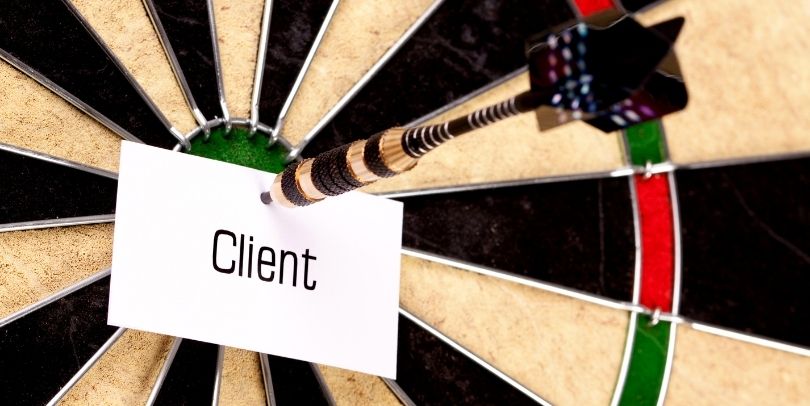 A dart is holding a piece of paper, with the word "Client" on it, in the bulls eye area of a dart board.